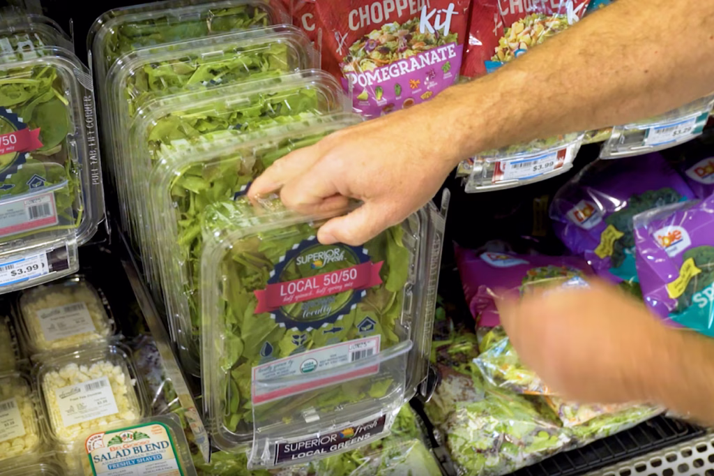 Hand pulling a clamshell of salad greens from a refrigerated produce shelf
